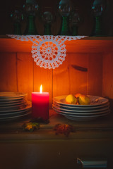 View into an open doors of a vintage cupboard illuminated with a lit red candle in a dark room, with old dishes, apples and crocheted doily. Autumn composition or happy holiday season background.