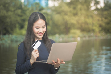 Young beautiful Asian woman using credit card and computer laptop for online shoping at outdoor garden park.