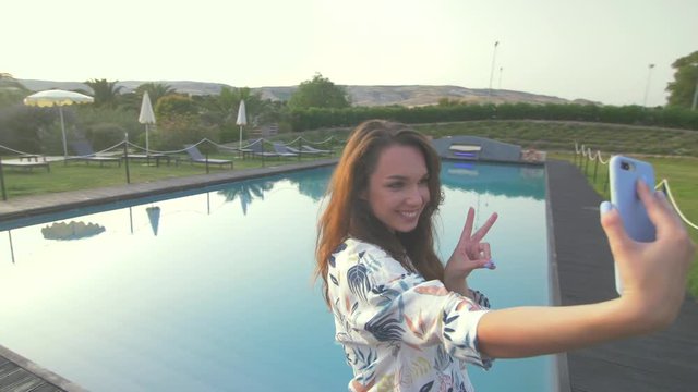 Beautiful woman making selfie mobile photo on nice luxury villa and swimming pool. Young girl taking photo.Female tourist at sunset.Elegant girl have good moments. Say hello and giving air kiss.