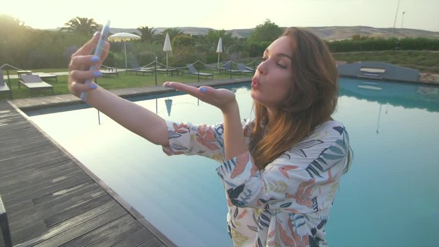Beautiful woman making selfie mobile photo on nice luxury villa and swimming pool. Young girl taking photo.Female tourist at sunset.Elegant girl have good moments. Say hello and giving air kiss. 