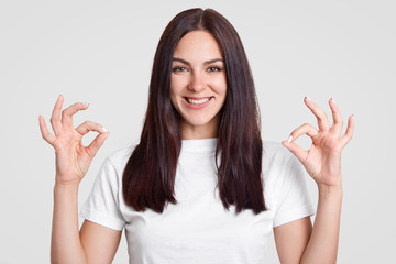 Indoor shot of happy attractive woman with long straight dark hair, makes okay sign with both hands, shows approval, dressed in casual outfit, poses against white background. Body language concept