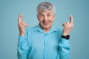 Horizontal shot of wrinkled man in blue formal shirt crosses fingers for good luck, being on pension, wears smartwatch, poses against blue background. Elderly people and body language concept