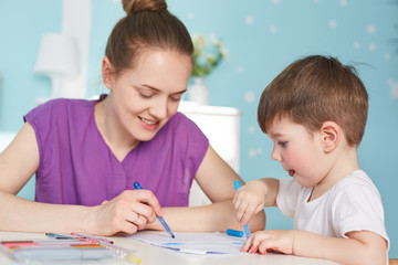 Photo of affectionate mother draws picture to her little son, spend ime together at home, have positive smiles, friendly relationships. Young mum takes care of child, gives educational lesson