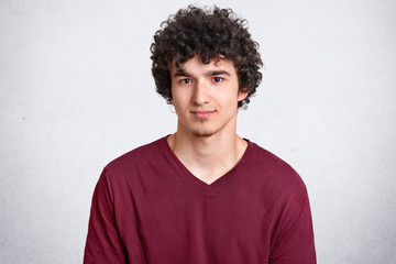 Fototapeta na wymiar Headshot of attractive curly youngster looks seriously at camera, has appealing look, spends free time with friends, dressed in casual outfit, isolated over white concrete wall. Youth concept