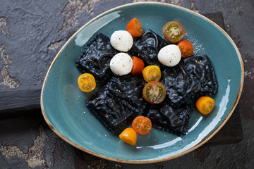 Black ravioli with cherry tomatoes and mozzarella served on a turquoise plate, high angle view, horizontal shot