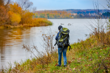 a tourist with a backpack on his shoulders walks along the river on a cold autumn day. A man dressed in warm clothes examines the area around