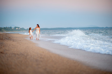happy mother walking with her beloved daughter on the beach at sunset, holding hands and smiling