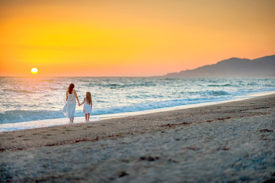 Mom and daughter in long white dresses walking to the beach on a beautiful sunset