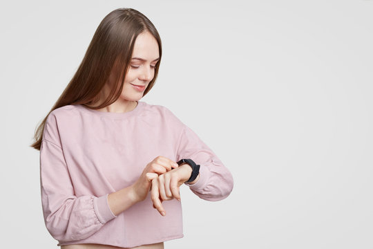Attractive woman looks at smartwatch, checks calories or pulse, wears casual oversized sweater, has dark staight hair, isolated on white background with free space. Modern technologies and innovations