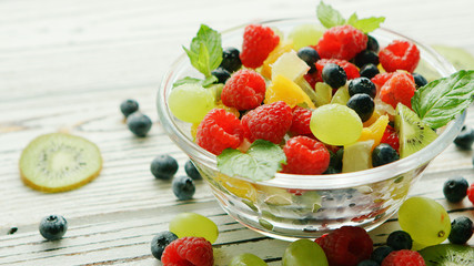 Glass bowl filled with sweet assortment of fruit and berries garnished with mint leaves and served on wood table