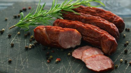 Closeup shot of delectable smoked sausages lying near twig of aromatic rosemary and seeds of hot pepper on scratched cutting board