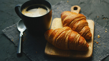 From above view of two fresh croissants and black mug with coffee placed on napkin on gray background of table