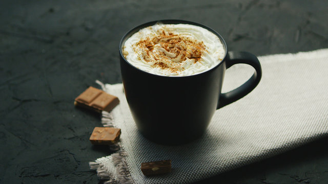 From above view of black mug of coffee with whipped cream on top and pieces of chocolate laid near on gray background