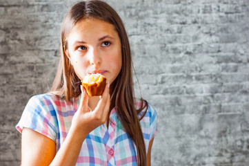 portrait of young teenager brunette girl with long hair eating muffin cake on gray wall background