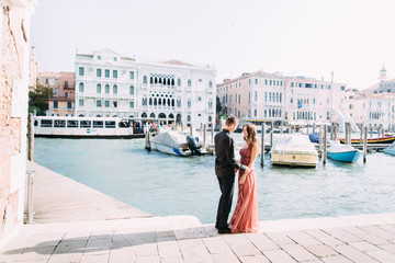 Fototapeta na wymiar Romantic photo of hugging and kissing couple in Venice. Young couple, girl in pink dress and boy in black clothes standing on the background of wonderful Venice buildings. Italy, Europe.