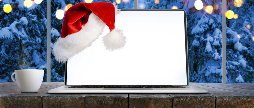 Laptop on table with Santa Claus hat at home with panoramic view through window of snowy trees in winter forest, screen is cut with provided clipping path