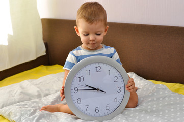 Cute toddler boy holding a big round clock sitting on the bed before going to bed