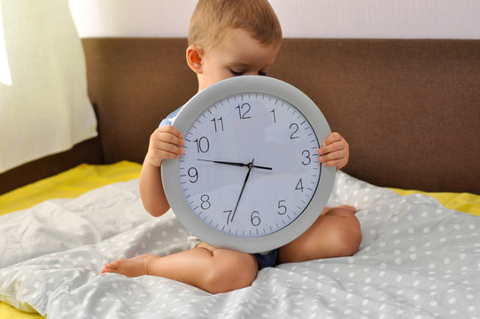 Cute Toddler Boy Holding A Big Round Clock Sitting On The Bed Before Going To Bed