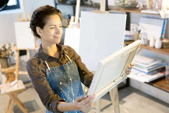 Happy woman in workwear looking at picture that she painted while holding it in hands