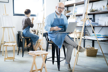 Fototapeta na wymiar Serious aged man in workwear using modern gadgets while working in studio of arts