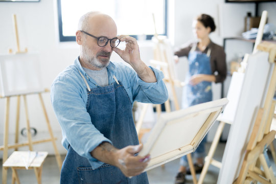 Serious man in eyeglasses and workwear looking at painting in his hands while standing by easel in studio