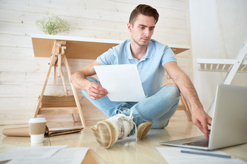 Young economist with paper sitting on the floor and searching for online data in laptop