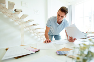 Young businessman reading one of financial papers while leaning over desk in modern studio