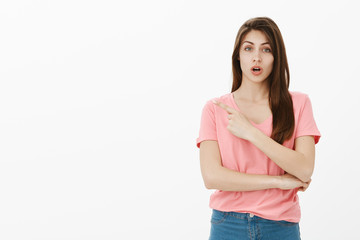 Girl asking advice while pointing at dress she wants to buy. Portrait of good-looking busy young woman in casual outfit, pointing at upper left corner and talking to camera over grey wall