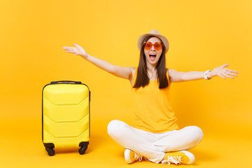 Fun tourist woman in summer casual clothes, hat sit with suitcase, spreading hands isolated on yellow orange background. Girl traveling abroad to travel on weekends getaway. Air flight journey concept