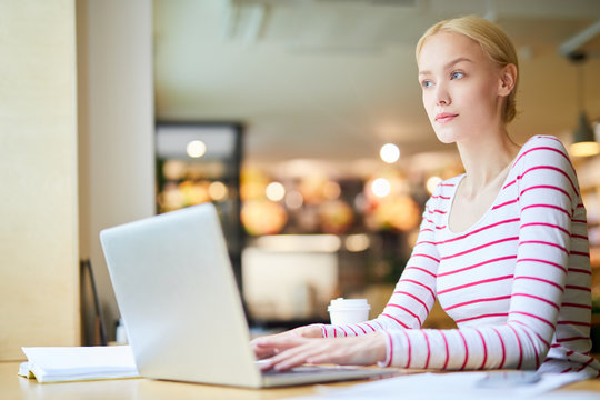 Young pensive blond woman in striped pullover sitting in cafe in front of laptop and carrying out task