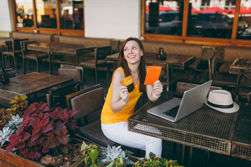 Woman in outdoors street cafe sitting at table with modern laptop pc computer, hold in hand bank credit card, passport, book online airplane ticket. Restaurant in free time. Lifestyle business concept