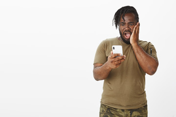 Amazed overwhelmed attractive plump guy in military clothes, holding smartphone, dropping jaw and...