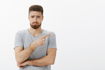 Sad and cute charming bearded male model in grey t-shirt frowning making gloomy face with frowned eyebrows, sulking pointing right expressing regret and envy standing unhappy over white background