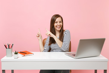 Young laughing woman in casual clothes point index finger aside sit, work at white desk with...