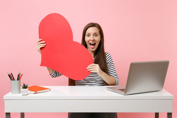 Young excited amazed woman holding red empty blank heart sit, work at white desk with pc laptop...