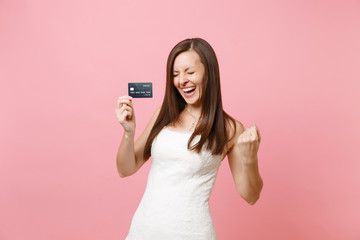 Joyful bride woman in white wedding dress holding credit card and doing winner gesture isolated on pink pastel background. Organization of wedding celebration concept. Copy space for advertisement.