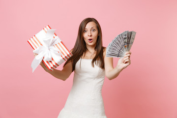 Amazed shocked bride woman in white wedding dress holding bundle lots of dollars, cash money, red box with gift, present isolated on pink pastel background. Wedding celebration concept. Copy space.