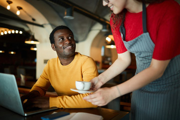 African-american guy looking at young waitress bringing him cup of tea or coffee