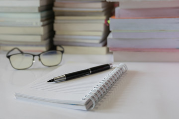Close up of notebook with ink-pen and eyeglasses put on white table with blur focus of books as a background. Educational concept.