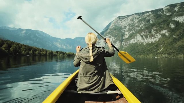 Girl traveler swims in boat on mountain lake surrounded by high mountains. Girl in canoe rowing leisurely paddle along calm lake. Woman in the boat from the back.