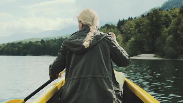 Girl traveler swims in boat on mountain lake surrounded by high mountains. Girl in canoe rowing leisurely paddle along calm lake. Woman in the boat from the back.