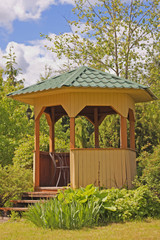 Gazebo in a landscaped garden surrounded by different plants. Yellow wooden gazebo with a green...