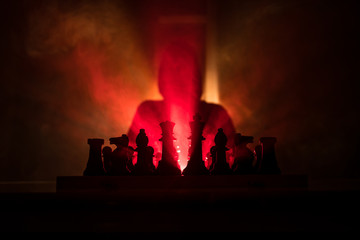 Fototapeta premium Man playing chess. Scary blurred silhouette of a person at the chessboard with chess figures. Dark toned foggy background.