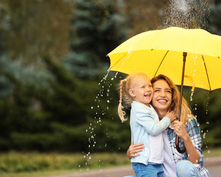 Happy mother and daughter with umbrella under rain in park. Space for text