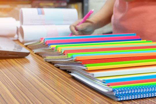 Asian teacher is checking pile of student's homework assignments on table which has a large number of paperworks stacked in archive with colorful plastic binding bars. Education and business concept.