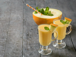Two glass mugs with melon smoothie and a half melon on a black wooden table.