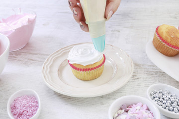 Woman prepares muffins decorated with cream and sprinkles.