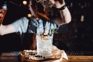 Barman prepares cocktail with orange and herbs in transparent glass on bar with alcohol. Uses burner with sparks. Dark background. - obrazy, fototapety, plakaty