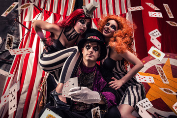 Three people performing on stage freak show, showing magic tricks with cards on dark circus scene