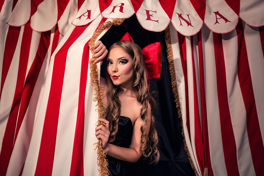 Beautiful and sexy with doll face with red bow and red lips in black dress peeps out with attractive look from behind the scenes of dark circus arena
